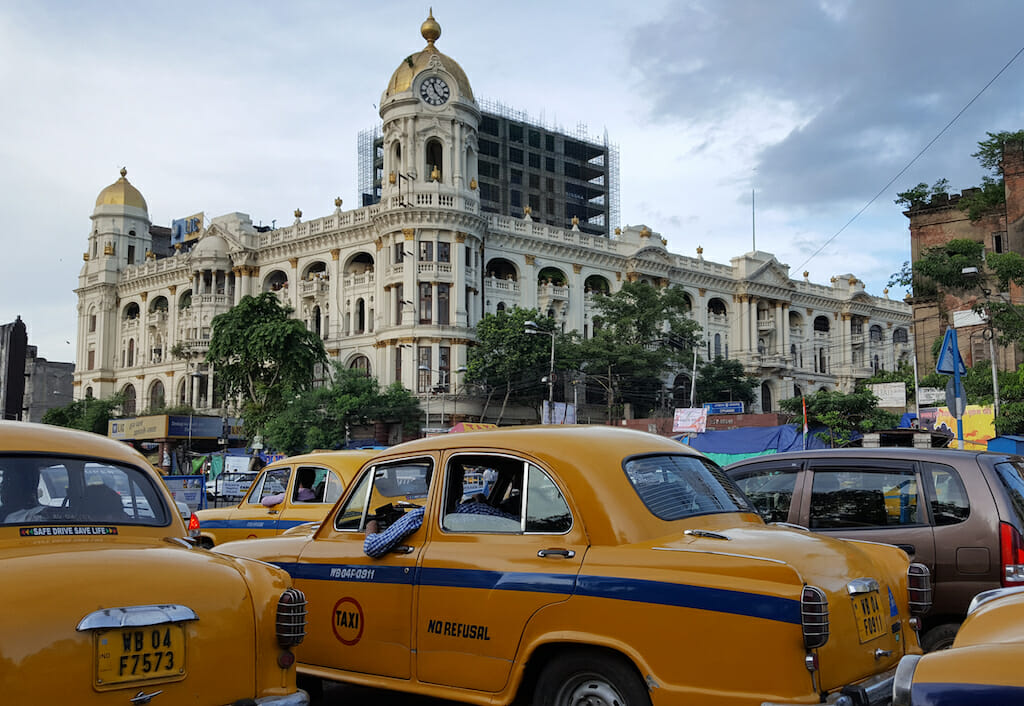 Yellow cabs flooding the streets along with other cars with a white historical building in the back on a sunny Kolkata day
