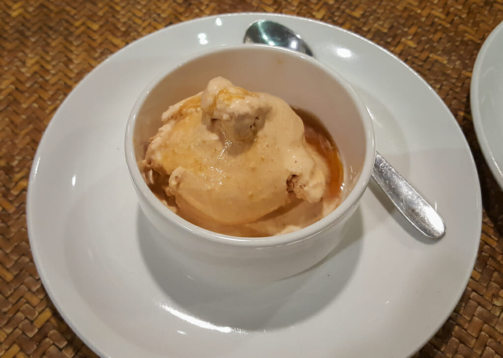 A little white bowl filled with caramel colored ice cream on a white saucer with a silver spoon next to it
